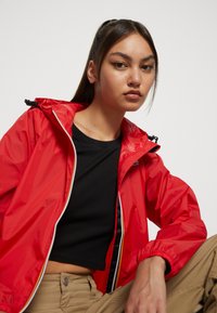 Chaqueta roja, ligera y impermeable con cremallera frontal y capucha ajustable. Combinada con una blusa negra de canalé y pantalones beiges.