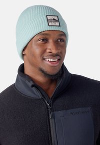 Gorro de punto verde menta con textura acanalada y una etiqueta negra con texto blanco, usado con una chaqueta de forro polar negra con cremallera.