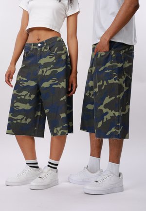 Dwie osoby stojące obok siebie, ubrane w białe trampki i krótkie spodnie w kamuflażowym wzorze do kolan, jedna z nich ma na sobie krótki biały top.