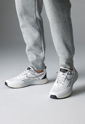 Person trägt graue Jogginghosen und weiße Sportschuhe mit schwarzen Sohlen und steht auf einem grauen Boden.