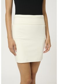 Femme portant une mini-jupe ivoire taille haute associée à un top noir sans manches, debout les bras détendus le long du corps.