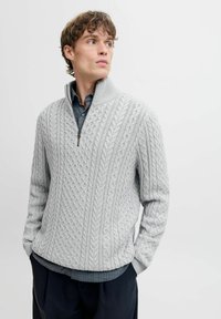 Hellgrauer Pullover mit hohem Kragen, ausgestattet mit einem Zopfmuster, Halbzippverschluss und gerippten Bündchen, über einem dunkelblauen Hemd getragen.