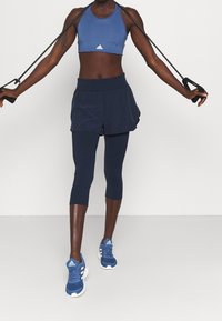 Námořnický sportovní outfit zahrnující modrý crop top, vrstvené šortky a legíny do kolen. Doplňky zahrnují cvičební gumy a modré tenisky.