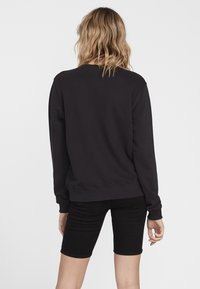 Mustat crewneck-hupparit pitkillä hihoilla, pehmeää kangasta, ribbikuvioidut hihansuut ja helma; yhdistettynä mustiin farkkushortseihin. Näkyvä ompelutaito.
