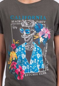 T-shirt gris avec un graphique coloré représentant un squelette portant des lunettes de soleil, vêtu d'une chemise florale bleue et entouré de fleurs tropicales.