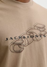 Beżowa koszulka bawełniana z graficznym wzorem w ciemnym brązie, z napisem "JACK & JONES" oraz warstwowymi kształtami geometrycznymi. Krótkie rękawy.