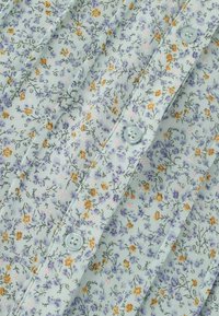 Zbliżenie jasnoniebieskiej tkaniny z małym fiołkowo-pomarańczowym wzorem kwiatowym oraz niebieskimi guzikami na plisowanej części ubrania.