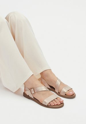 Nohy v béžových kožených sandálech se dvěma pásky a přezkou, doplněné volnými krémovými kalhotami na bílém pozadí.