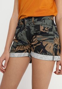 Pantalones cortos de camuflaje en verde, negro y marrón, con puños de denim enrollados y bolsillos frontales. Hechos de algodón con un ajuste estructurado.