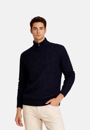Jeune homme portant un pull en maille torsadée bleu marine foncé avec une fermeture éclair quart de zip et un pantalon beige clair, debout avec une main dans la poche.