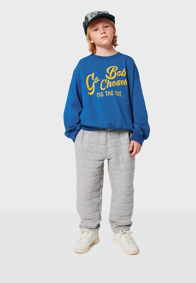 Felpa blu con testo grafico giallo, pantaloni grigi trapuntati e sneaker bianche. L'outfit presenta un colletto rotondo e una vestibilità comoda.