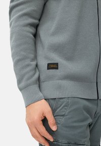 Grijze gebreide trui met een gestructureerde afwerking, geribbelde manchetten en een stoffen label met de tekst "Made for daily adventures" in zwart en goud.