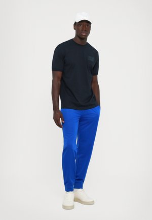 Man wearing a black t-shirt, bright blue jogger pants, white sneakers, and a white cap standing with hands in pockets on a white background.