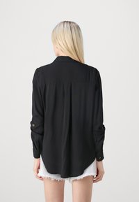 Camisa negra de manga larga con cuello, textura suave y mangas remangadas. Presenta detalles de botones y un dobladillo ligeramente curvado en la parte de atrás.