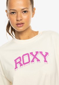Camiseta color crema con el texto "ROXY" en neón rosa, en un estilo audaz y estilizado. Material suave con un corte relajado y escote redondo.