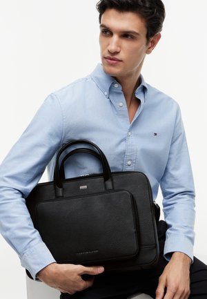 Young man in light blue button-up shirt holding a black leather Tommy Hilfiger briefcase against a plain white background.