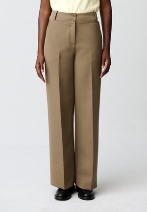 Persona che indossa pantaloni beige a vita alta e gamba larga con pieghe frontali e scarpe nere, in piedi davanti a uno sfondo chiaro e uniforme.