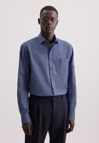 Uomo che indossa una camicia button-up blu con punti geometrici bianchi e pantaloni neri eleganti, in piedi contro uno sfondo chiaro e uniforme.