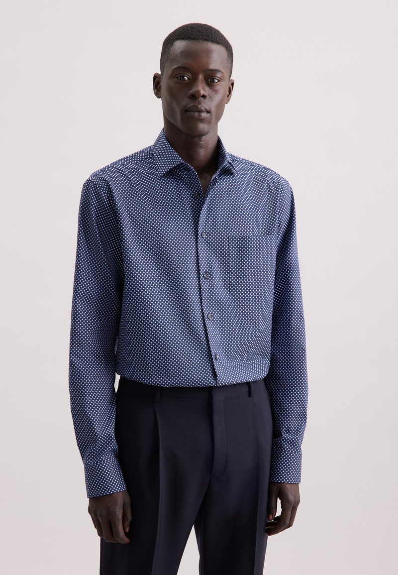 Uomo che indossa una camicia button-up blu con punti geometrici bianchi e pantaloni neri eleganti, in piedi contro uno sfondo chiaro e uniforme.