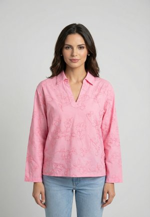 Woman wearing a long-sleeve pink blouse with subtle floral embroidery and light blue jeans, standing against a plain light gray background.