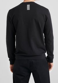 Sudadera negra de manga larga hecha de un tejido suave, con un cuello acanalado y un patrón de rayas blancas en la parte superior de la espalda. Textura suave.