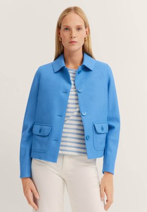 Blaues Wolljackett mit kragenförmigem Ausschnitt, zwei Fronttaschen und Knopfverschluss. Getragen über einem gestreiften Hemd und kombiniert mit weißen Hosen.