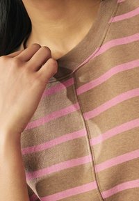 T-shirt marrone con strisce orizzontali rosa, con scollatura rotonda e tessuto testurizzato. Mano che tocca delicatamente il colletto.