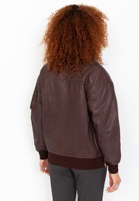 Veste bombardier en cuir marron avec des poignets et une taille côtelés, dotée d'une poche zippée sur la manche, texture lisse et coupe décontractée.