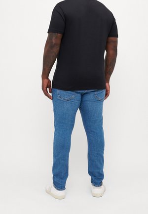 Person trägt ein schwarzes kurzärmeliges Shirt, blaue Jeans und weiße Sneakers und steht mit dem Rücken zur Kamera vor einem einfarbigen Hintergrund.