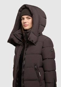 Giacca a vescica marrone con colletto alto, grande cappuccio, chiusure con zip e bottoni a pressione, dotata di tasche laterali e finitura opaca.