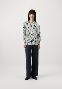 Hellgraue Bluse mit schwarzen abstrakten Mustern, durchgehender Knopfleiste und langen Ärmeln. Kombiniert mit dunklen, weit geschnittenen Hosen und schwarzen Schuhen.