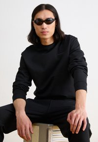 Felpa nera con polsini e colletto a costine, caratterizzata da un logo discreto. Abbinata a pantaloni neri. Occhiali da sole eleganti completano il look.