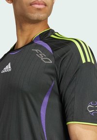 Zwart sportshirt met ribbeltextuur, voorzien van paarse en groene accenten, drie groene strepen op de schouders en een wit Adidas-logo.