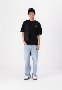 T-shirt en coton noir avec un texte blanc, jeans en denim bleu clair et sac en bandoulière noir. Des baskets grises et blanches complètent la tenue.