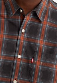 Camisa de cuadros en negro, naranja y azul. Presenta un cuello clásico, botones blancos, y un bolsillo en el pecho con una etiqueta de logo roja.