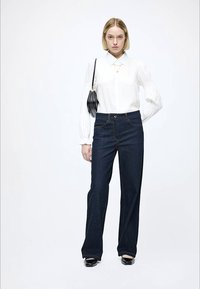 Tumši zila džinsa plata piegriezuma bikses kopā ar baltu blūzi ar apkakli. Modele tur rokā nelielu melnu somiņu. Gluda tekstūra un pielāgots piegriezums.