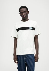 Svijetlosivi T-shirt s crnom horizontalnom prugom, s plavom logotipom. Mekani pamučni materijal, opušteni kroj, kratki rukavi.