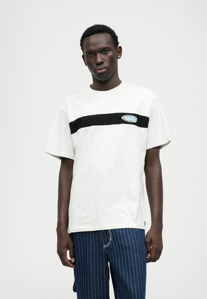 Lys grå T-shirt med en sort vandret stripe, der har et blåt logo-patch. Blødt bomuldsstof, afslappet pasform, korte ærmer.