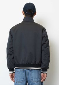 Giacca bomber nera con polsini e orlo a coste, caratterizzata da una texture liscia e un colletto alla coreana. Indossata con jeans in denim azzurro.