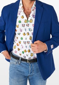 Blazer blu sopra una camicia bianca a maniche lunghe con personaggi pixelati colorati. Indossato con jeans in denim azzurro chiaro e una cintura nera texturizzata.
