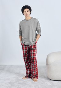 Gray long-sleeve shirt with relaxed fit paired with red plaid pajama pants featuring black and white accents. Model stands barefoot on gray carpet.