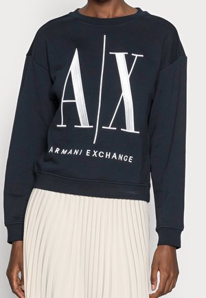 Persona que lleva una sudadera negra de manga larga de Armani Exchange con un gran logo "A|X" y una falda plisada blanca visible.