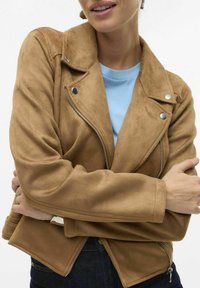 Veste en faux daim marron de style biker avec des zips argentés et des boutons-pression, présentant un design ajusté et des revers larges. T-shirt bleu en dessous.