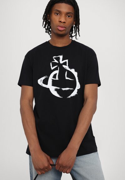 Tricou negru din bumbac, cu un design grafic alb mare care prezintă un motiv cu cruce și planete. Modelul ține curelele, purtând blugi deschiși la culoare.