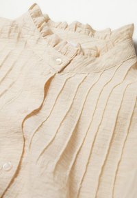 Blouse texturée couleur crème avec col volanté, plis verticaux et petits boutons ronds le long de la patte de boutonnage avant.