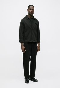 Mustast puuvillast jakk krae ja nuppudega, kantud musta särgi üle, kombineeritud mustade sirgete pükste ja mustade tossudega.