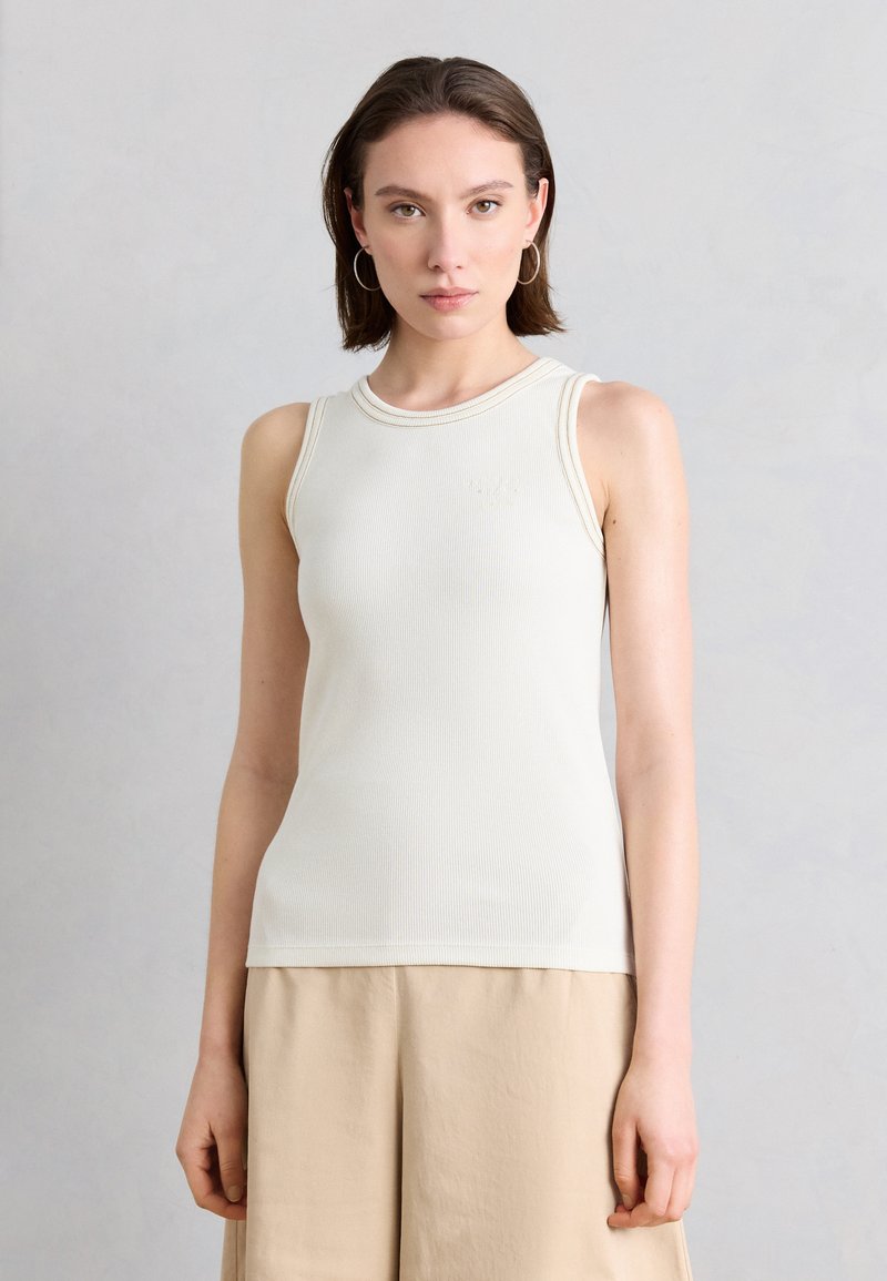 Γυναίκα με κοντά καστανά μαλλιά που φοράει ασημένια σκουλαρίκια κρίκους, λευκή αμάνικη ριγωτή μπλούζα και μπεζ παντελόνι, στέκεται μπροστά σε ανοιχτό γκρι φόντο.