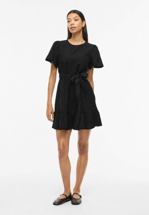 Kvinde står iført en kort sort kjole med pufærmer, et bindebælte i taljen og sorte flade sko mod en ensartet hvid baggrund.