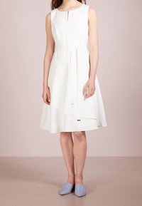 Robe blanche sans manches avec un décolleté en trou de serrure, taille cintrée par un lien, et un ourlet superposé. Associée à des chaussures légères bleu clair à enfiler.