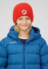 Giovane con capelli biondi lunghi che indossa un berretto rosso con un logo bianco di un mammut e una giacca imbottita blu Mammut, sorridendo dolcemente.